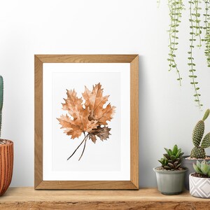 May include: A framed watercolour painting of three brown oak leaves on a white background. The frame is made of light brown wood and is sitting on a wooden shelf with a cactus, succulents, and hanging plants in the background.