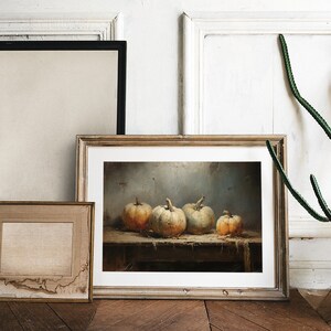May include: A framed painting of a still life with four pumpkins on a wooden table. The pumpkins are various shades of orange and white. The painting is in a light wood frame and is hanging on a white wall.