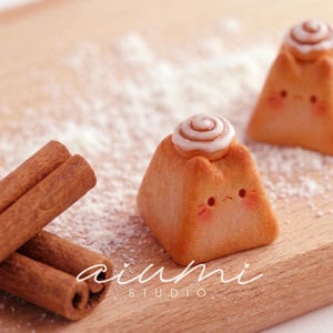 May include: Two small, tan, pyramid-shaped figurines with cat-like ears and pink cheeks, each topped with a white-frosted cinnamon roll. The figurines are placed near two brown cinnamon sticks and a dusting of white powder.