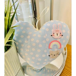 May include: A blue heart-shaped cushion with white polka dots and embroidered animals. The cushion features a pink bunny, a rainbow, and a cat.