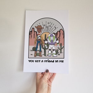 May include: A print featuring a cartoon illustration of Woody and Buzz Lightyear from Toy Story standing in a desert landscape with the text "You got a friend in me".
