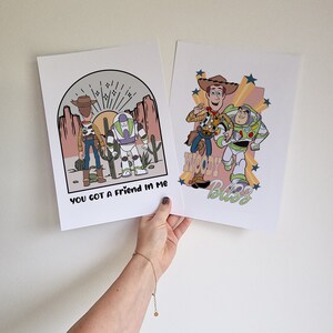 May include: Two prints featuring illustrations of Woody and Buzz Lightyear from the Toy Story franchise. The first print shows Woody and Buzz standing together in a desert landscape with the text "You got a friend in me". The second print shows Woody and Buzz running together with the text "Woody's Buzz".