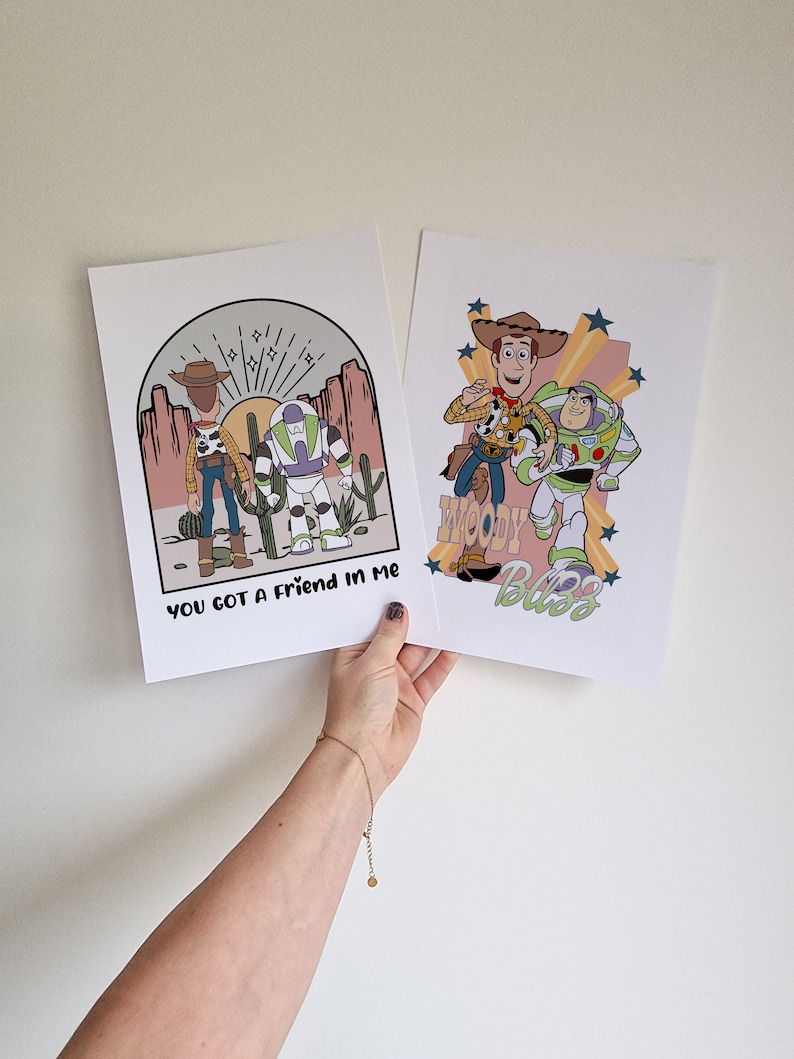 May include: Two prints featuring illustrations of Woody and Buzz Lightyear from the Toy Story franchise. The first print shows Woody and Buzz standing together in a desert landscape with the text "You got a friend in me". The second print shows Woody and Buzz running together with the text "Woody's Buzz".
