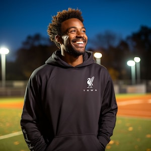 Puede incluir: Una sudadera con capucha negra con un escudo blanco del Liverpool Football Club y el texto "YNWA" en la parte delantera.