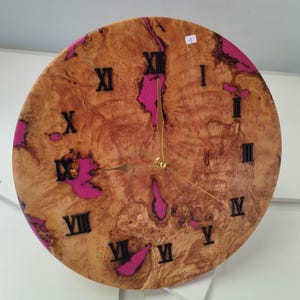 Può includere: Orologio rotondo in legno con un motivo a venature naturali e accenti in resina rosa. Il quadrante presenta numeri romani neri e lancette dorate. Un oggetto decorativo.