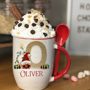 May include: White ceramic mug with a red handle and a spoon attached. The mug features a gold glitter letter O with a Christmas gnome and presents around it. The mug says "Leave it with me" repeatedly around the side. The mug also has the name "Oliver" written in red. The mug is filled with whipped cream, chocolate chips, and chopped nuts. There is a chocolate bar sticking out of the whipped cream.