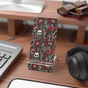 Puede incluir: Un soporte rectangular para teléfono con fondo gris oscuro y un patrón repetitivo de calaveras blancas, labios de vampiro rojos con colmillos, frascos de pociones rojos, ataúdes blancos, rosas rojas y pequeños murciélagos negros.