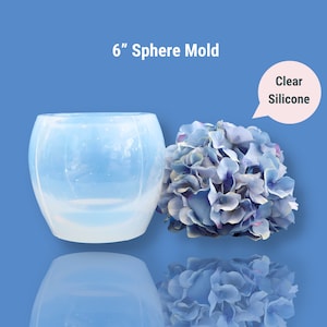 May include: A clear silicone sphere mould, approximately 15 cm in diameter, next to a cluster of blue and purple artificial flowers. The mould has a rounded shape and is made of clear material. Text on the image reads "6" Sphere Mold" and "Clear Silicone."
