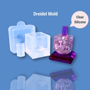 Könnte beinhalten: Transparente Silikonform zum Herstellen eines Dreidels. Die Form umfasst eine quadratische Basis, eine runde Oberseite und eine separate Form für den Dreidel-Stiel. Die Form wird mit einem fertigen Dreidel aus klarem Harz und rosa Glitzer gezeigt.