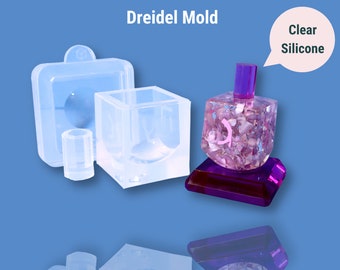 May include: Clear silicone mold for making a dreidel. The mold includes a square base, a round top, and a separate mold for the dreidel's stem. The mold is shown with a finished dreidel made with clear resin and pink glitter.