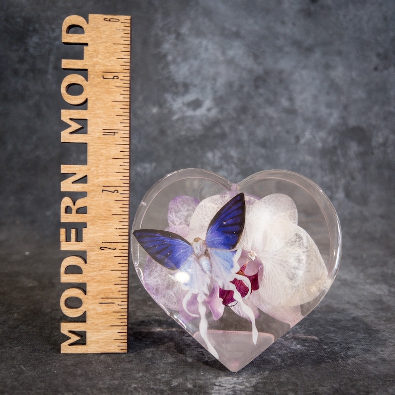 K&ouml;nnte beinhalten: Eine transparente, herzf&ouml;rmige Harzform mit einem blauen Schmetterling und lila und wei&szlig;en Blumen. Ein Holzlineal mit dem Text "MODERN MOLD" befindet sich links von der Form. Die Form ist etwa 7.6 cm breit.