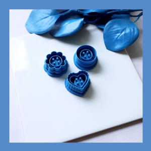 Peut inclure: Trois emporte-pièces bleus en forme de fleur, de cercle et de cœur. Les emporte-pièces sont en plastique et sont disposés sur une surface blanche avec des feuilles décoratives bleues en arrière-plan. Les emporte-pièces sont pour la pâtisserie.