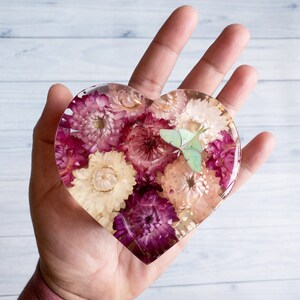 K&ouml;nnte beinhalten: Herzf&ouml;rmiges Kunstwerk aus Harz, gef&uuml;llt mit getrockneten Blumen in Rosa, Lila und Creme. Ein hellgr&uuml;ner Schmetterling ist in das Harz eingebettet. Das St&uuml;ck wird in einer Hand gehalten und zeigt das klare Harz und die Blumenanordnung.