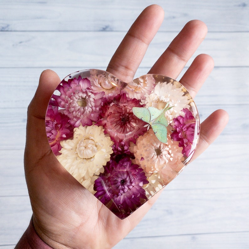 K&ouml;nnte beinhalten: Herzf&ouml;rmiges Kunstwerk aus Harz, gef&uuml;llt mit getrockneten Blumen in Rosa, Lila und Creme. Ein hellgr&uuml;ner Schmetterling ist in das Harz eingebettet. Das St&uuml;ck wird in einer Hand gehalten und zeigt das klare Harz und die Blumenanordnung.