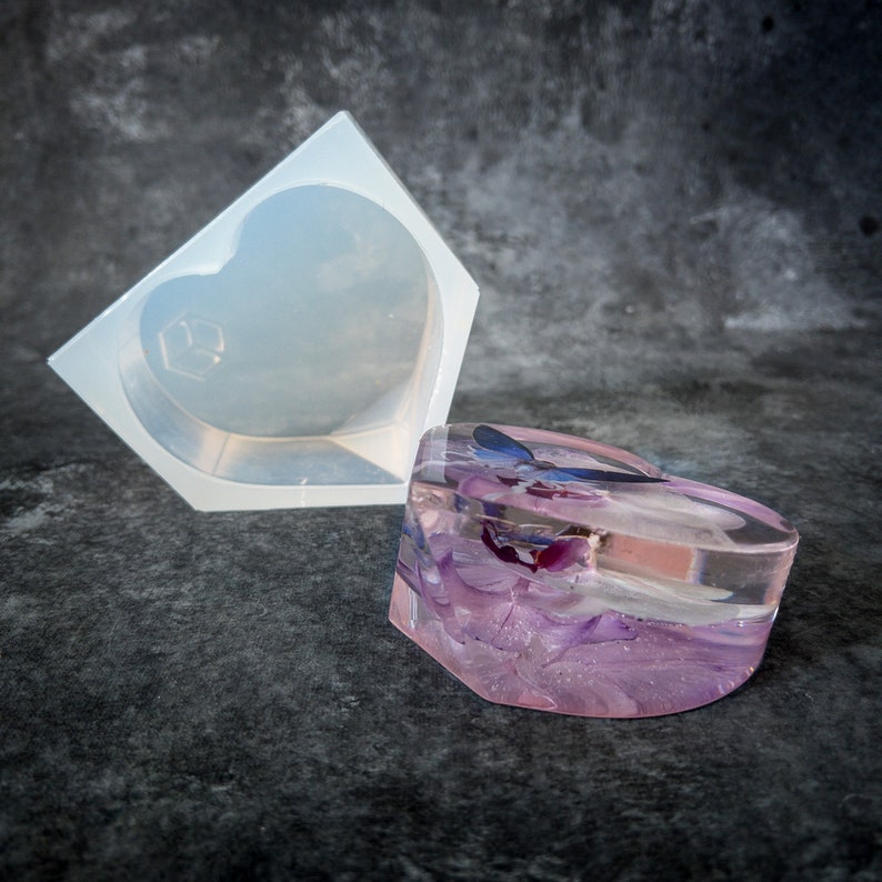 K&ouml;nnte beinhalten: Eine herzf&ouml;rmige Harzbox mit Deckel, gef&uuml;llt mit lila und blauen Trockenblumen. Daneben befindet sich eine transparente Silikonform in Herzform. Die Box hat einen rosafarbenen Farbton und steht vor einem dunkelgrauen Hintergrund.