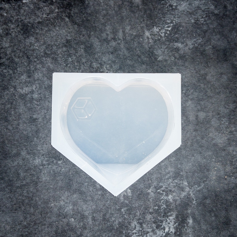 K&ouml;nnte beinhalten: Eine klare, herzf&ouml;rmige Silikonform mit einem geometrischen W&uuml;rfeldesign. Die Form ist in einem wei&szlig;en, eckigen Rahmen angeordnet, ideal f&uuml;r die Herstellung von Kunstharz oder anderen DIY-Projekten. Die Form befindet sich auf einer dunkelgrauen, strukturierten Oberfl&auml;che.