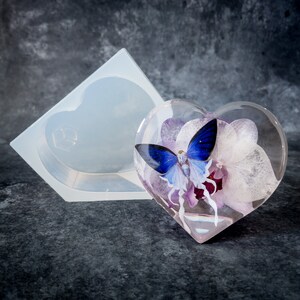 K&ouml;nnte beinhalten: Ein klarer, herzf&ouml;rmiger Papierbeschwerer aus Harz mit einem blauen Schmetterling und lila Blumen im Inneren. Eine Silikonform in Herzform befindet sich neben dem Papierbeschwerer. Der Hintergrund ist eine dunkelgraue Oberfl&auml;che.
