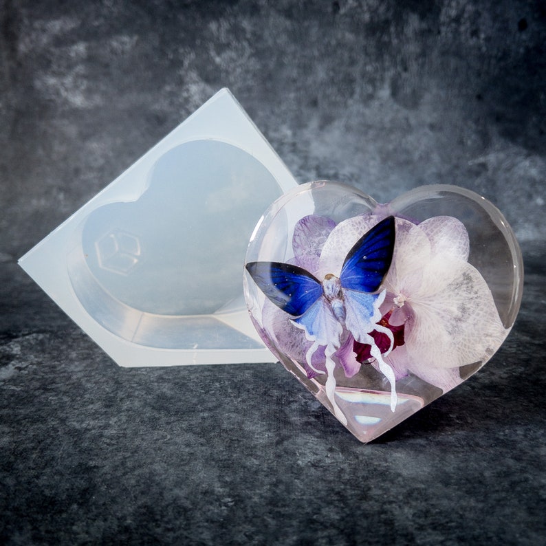 K&ouml;nnte beinhalten: Ein klarer, herzf&ouml;rmiger Papierbeschwerer aus Harz mit einem blauen Schmetterling und lila Blumen im Inneren. Eine Silikonform in Herzform befindet sich neben dem Papierbeschwerer. Der Hintergrund ist eine dunkelgraue Oberfl&auml;che.