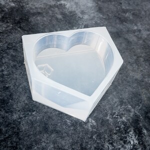 K&ouml;nnte beinhalten: Eine transparente Silikonform in Herzform mit rechteckiger Basis. Die Form ist klar und liegt auf einer dunklen, strukturierten Oberfl&auml;che. Die Herzform ist deutlich im Inneren der Form zu sehen.