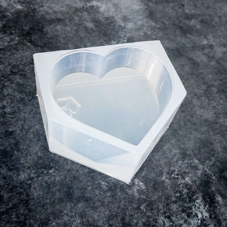 K&ouml;nnte beinhalten: Eine transparente Silikonform in Herzform mit rechteckiger Basis. Die Form ist klar und liegt auf einer dunklen, strukturierten Oberfl&auml;che. Die Herzform ist deutlich im Inneren der Form zu sehen.