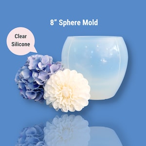 Pode incluir: Uma forma de esfera de silicone transparente de 20,3 cm para artesanato. A forma está sobre uma superfície azul com duas flores artificiais ao lado.