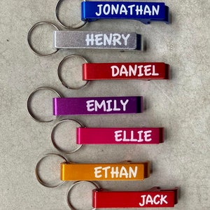 May include: Nine personalized bottle openers with key rings. The bottle openers are in different colors, including purple, blue, silver, red, pink, orange, and black. Each bottle opener has a name printed on it in white letters: Jessica, Jonathan, Henry, Daniel, Emily, Ellie, Ethan, Jack, and Kevin.