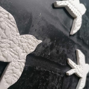 May include: White ceramic bird wall art with textured details. The birds are arranged in a dynamic composition against a dark, textured background, creating a sense of movement and flight. Decorative wall art.
