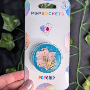 May include: A PopSocket phone grip with a blue background, featuring pink flowers and a gold-colored bee design. The PopSocket is on a white and iridescent backing with the text "POPSOCKETS" and "POPGRIP".