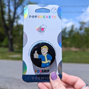 May include: A black and white PopGrip phone stand with a Fallout character giving a thumbs up. The text "Spooky Girl Designs" and "PopGrip" are printed on the stand. The stand is attached to a white and iridescent background with the word "POPSOCKETS" printed on it.