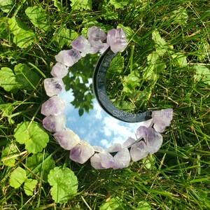 May include: A round mirror with a black frame, encircled by raw amethyst crystals. The mirror reflects the sky and is set against a backdrop of green grass and leaves. The text "SpookyGirlDesigns" is visible on the mirror.