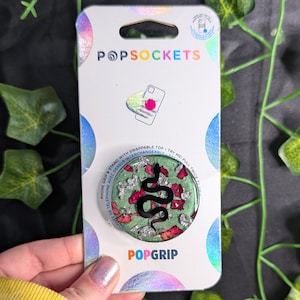 May include: A PopSockets PopGrip with a green background, a black snake design, and red and silver accents. The PopGrip is on a white and iridescent card with the PopSockets and PopGrip logos.