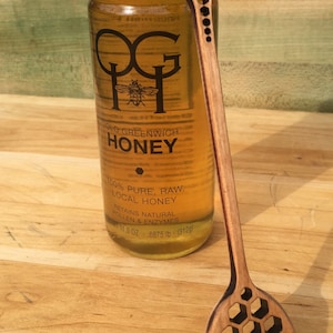 May include: A glass jar of honey with a black lid and a label that reads "Old Greenwich Honey" and "100% Pure, Raw, Local Honey". The jar is next to a wooden honey dipper with a bee design and a honeycomb pattern.