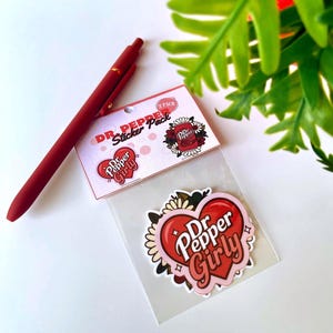 May include: A two-pack of Dr. Pepper themed stickers in a clear plastic sleeve with a header card. The stickers feature a heart shape with the text Dr. Pepper Girly and floral accents. A matte red retractable pen is positioned next to the sticker pack.