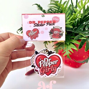 May include: A two-pack sticker set in a clear plastic sleeve with a header card labeled Dr. Pepper Sticker Pack. The stickers feature a red heart with the text Dr. Pepper Girly and a Dr. Pepper soda can surrounded by white and dark floral elements.