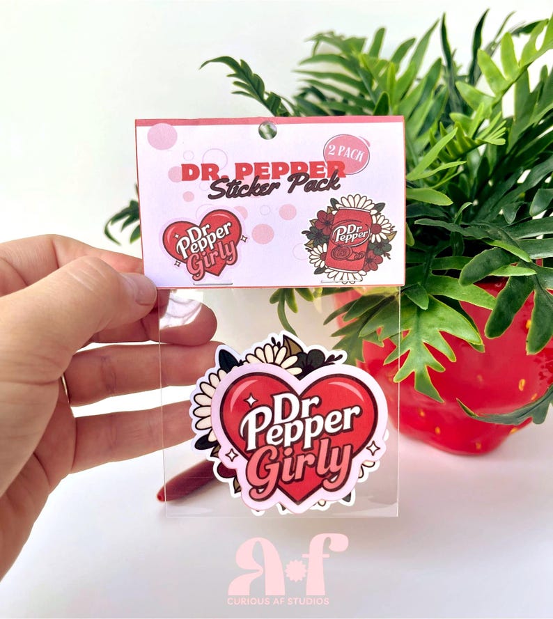 May include: A two-pack sticker set in a clear plastic sleeve with a header card labeled Dr. Pepper Sticker Pack. The stickers feature a red heart with the text Dr. Pepper Girly and a Dr. Pepper soda can surrounded by white and dark floral elements.