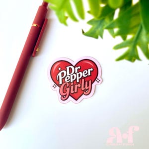 May include: A heart-shaped sticker featuring the text Dr Pepper Girly in white and pink lettering. The sticker has a red heart background with small white star accents. A matte red pen is positioned diagonally to the left of the sticker.