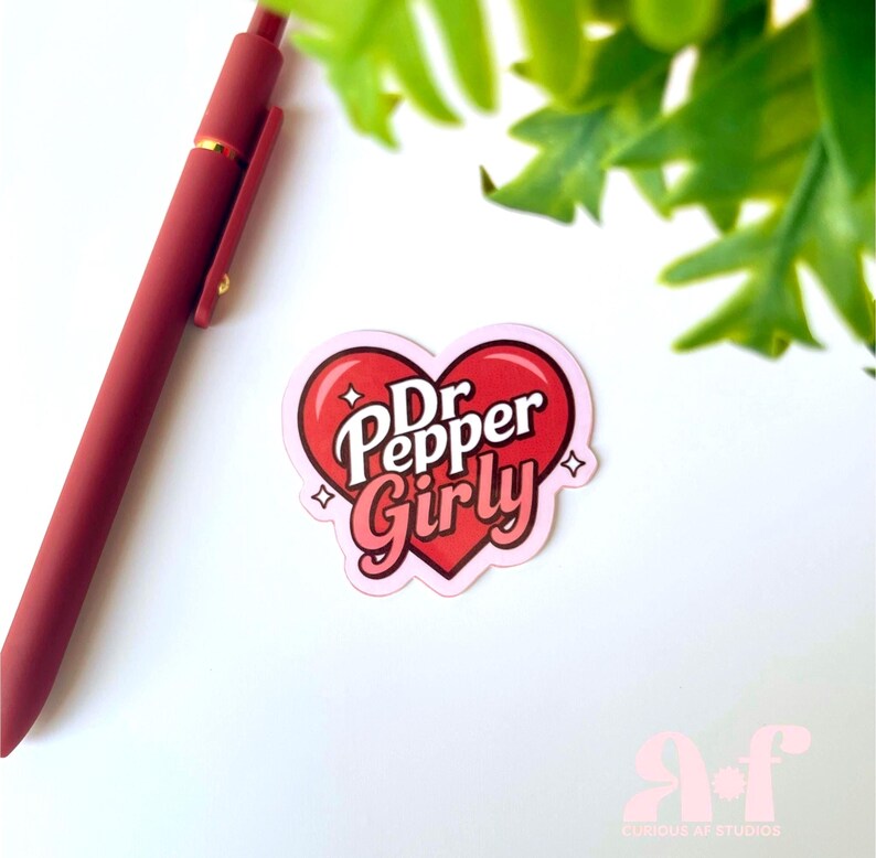 May include: A heart-shaped sticker featuring the text Dr Pepper Girly in white and pink lettering. The sticker has a red heart background with small white star accents. A matte red pen is positioned diagonally to the left of the sticker.