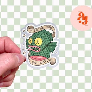 May include: A die-cut sticker featuring a green, scaly fish character with yellow eyes marked by black X's, sharp teeth, and pink lips. The fish is framed by two beige ribbons with the text FISHING FOR VALIDATION in black capital letters.