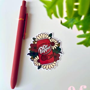 May include: A matte red retractable pen positioned next to a die-cut sticker featuring a red Dr. Pepper soda can surrounded by a cluster of cream, dark red, and dark green flowers. The sticker displays the Dr. Pepper logo and the number 23.
