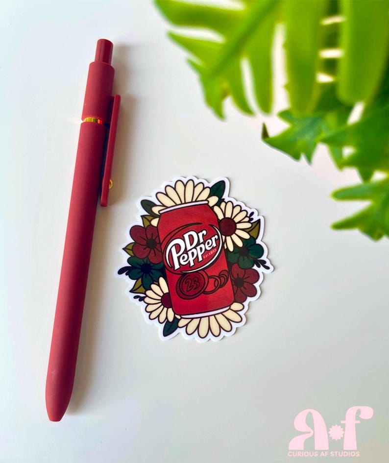 May include: A matte red retractable pen positioned next to a die-cut sticker featuring a red Dr. Pepper soda can surrounded by a cluster of cream, dark red, and dark green flowers. The sticker displays the Dr. Pepper logo and the number 23.