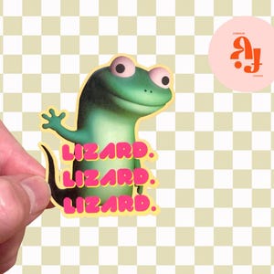 May include: A sticker featuring a cartoon lizard in shades of green and yellow, with the word "LIZARD" in pink repeated three times. The lizard is waving its arm. A pink circle with orange text is in the upper right corner.