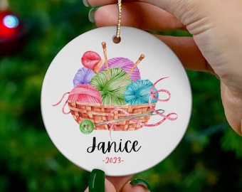May include: White ceramic Christmas ornament with a watercolor illustration of yarn balls in a basket. The ornament has the name "Janice" and the year "2023" written on it.