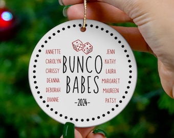 May include: A white ceramic ornament with the text "Bunco Babes - 2024" and the names of the group members. The ornament features a dice graphic and a gold string for hanging. The background is a Christmas tree with red ornaments.