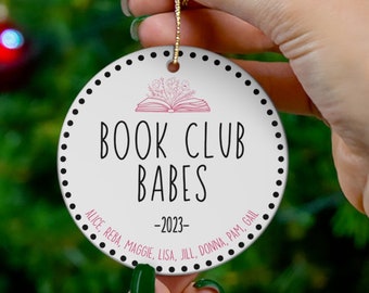May include: White round Christmas ornament with black polka dots and the text "Book Club Babes" in black with a pink book icon. The text "Alice, Reba, Maggie, Lisa, Jill, Donna, Pam, Gail - 2023" is written in black around the edge of the ornament.