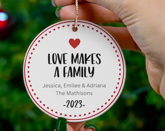 Puede incluir: Adorno navideño redondo blanco con lunares rojos y un corazón rojo. El adorno dice "Love Makes a Family" con los nombres "Jessica, Emilee & Adriana The Mathisons -2023- "