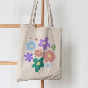 Puede incluir: Una bolsa de lona beige con un diseño floral. Las flores son en tonos de rosa, morado, azul y verde. La bolsa tiene dos asas y está colgada de un estante de madera.