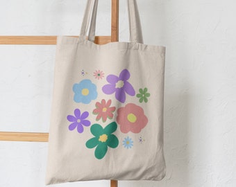 Peut inclure: Un sac fourre-tout en toile beige avec un motif floral. Les fleurs sont dans des tons de rose, violet, bleu et vert. Le sac a deux anses et est suspendu à un support en bois.