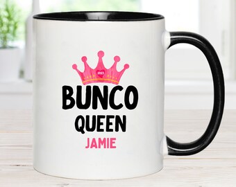 May include: White ceramic mug with a black rim and handle. The mug has a pink crown with the year 2023 on it and the text "Bunco Queen Jamie" in black.