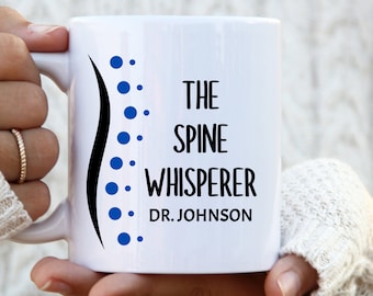May include: White ceramic mug with the text "THE SPINE WHISPERER DR. JOHNSON" in black. A black spine graphic with blue dots is on the left side of the mug. The mug is held by a person wearing a white sweater.