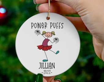 May include: White round ceramic ornament with a cartoon drawing of a cheerleader holding pom-poms. The text "Pomer Puffs" is at the top of the ornament, and the text "Jillian -2022- " is at the bottom.